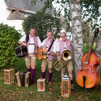 In the picture, three musicians are standing in traditional attire in a garden. All three are wearing lederhosen and light shirts and are posing with their instruments in front of several birch trees.
The left musician is holding a guitar, the middle one is also holding a guitar, and the right musician is holding a large brass instrument, presumably a tenor horn or baritone. To the right of the group, there is also a large double bass next to a tree.