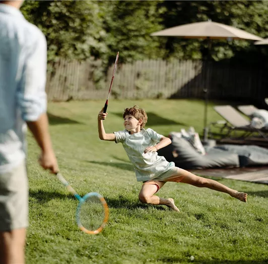 A child is playing badminton in the garden, in the background there are loungers and sun umbrellas visible.