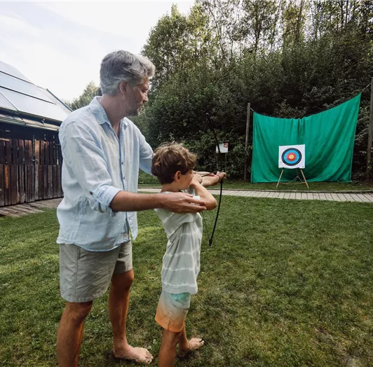 An adult is showing a child in the garden how to shoot with a bow. In the background, there is a target.