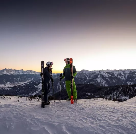 Zwei Skifahrer stehen auf einem schneebedeckten Berg und unterhalten sich. Im Hintergrund sind majestätische Berge und ein klarer Himmel zu sehen.
Letzte Abfahrt zum Apres Ski