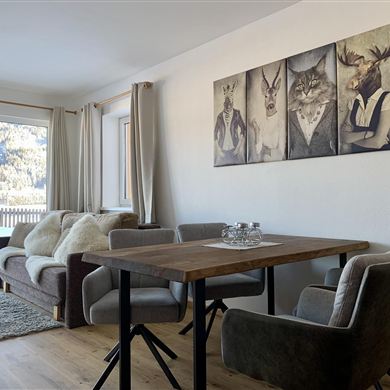 Cozy living experience with alpine flair. The stylishly furnished living area of our apartment combines modern design with rustic charm. Large windows offer a breathtaking view of the mountain landscape and provide plenty of daylight. The comfortable couch invites you to relax, while the spacious solid wood dining table offers enough space for sociable evenings. High-quality upholstery and warm wooden floors complete the cozy ambiance – ideal for restful days in the Alps.