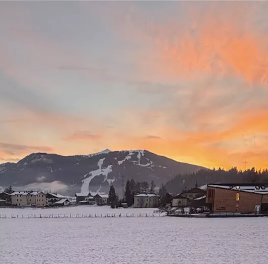 Eine winterliche Landschaft mit schneebedeckten Feldern und Bergen im Hintergrund. Der Himmel ist in warmen Farben bei Sonnenuntergang gefärbt.