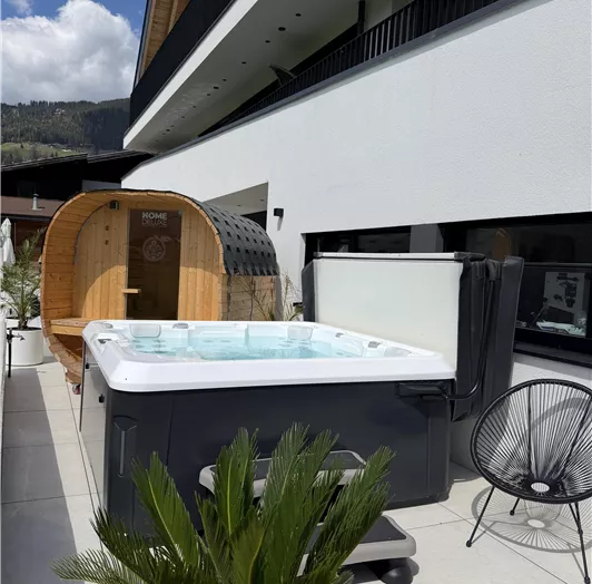 Ein moderner Outdoor Whirlpool und eine Outdoor Sauna auf einer Terrasse, umgeben von mediterranen Pflanzen. Im Hintergrund ist ein stilvolles, modernes Gebäude zu sehen.