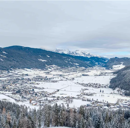 Eine schneebedeckte Landschaft mit einem kleinen Dorf im Tal. Umgeben von Bergen und Wäldern unter einem bewölkten Himmel.