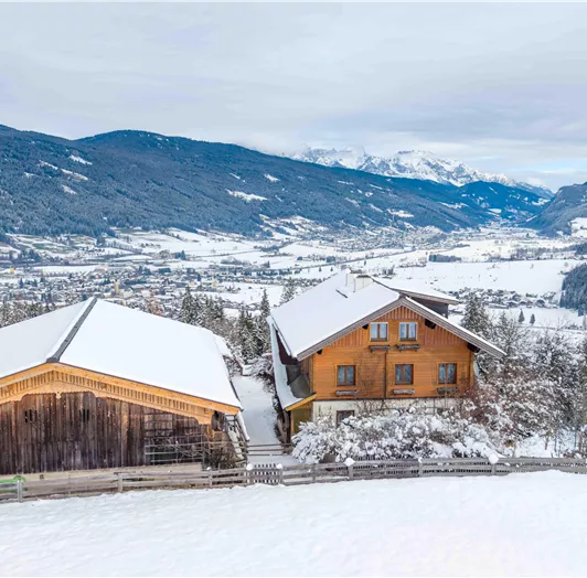 Eine winterliche Landschaft mit verschneiten Hügeln und Bergen im Hintergrund. Zwei gemütliche Holzhäuser stehen in der ruhigen Umgebung.