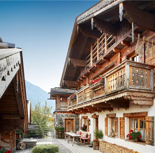 Authentische Chalet-Architektur mit Liebe zum Detail. Diese beeindruckende Außenansicht zeigt das Chalet in seiner ganzen Schönheit: Traditionelle Bauweise mit Naturstein, massivem Altholz und kunstvoll geschnitzten Holzbalkonen verleiht dem Haus seinen einzigartigen Charakter. Blumenkästen mit roten Geranien, Fensterläden mit Herzmotiven und dekorativ gestapeltes Kaminholz sorgen für ein einladendes und stimmungsvolles Ambiente. Eine gemütliche Sitzgelegenheit vor dem Haus rundet das harmonisch