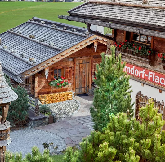 Einladender Hüttenzugang im Alpendorf Flachau. Der liebevoll gestaltete Weg zu den Chalets vermittelt auf den ersten Blick alpinen Charme und herzliche Gastlichkeit. Naturstein, Holzschindeln, traditionelle Fensterläden und blühende Blumenkästen prägen das authentische Erscheinungsbild. Der gepflegte Außenbereich fügt sich harmonisch in die umgebende Berglandschaft ein. Das große Schild verrät schon beim Ankommen: Willkommen im Almdorf Flachau – Ihrem Urlaubsort mit Herz.