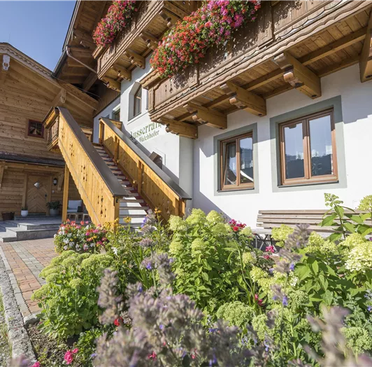 Ein traditionelles Haus mit Holzverkleidung und schönen Blumen im Vordergrund. Die Treppe führt zu einem gemütlichen Eingangsbereich.