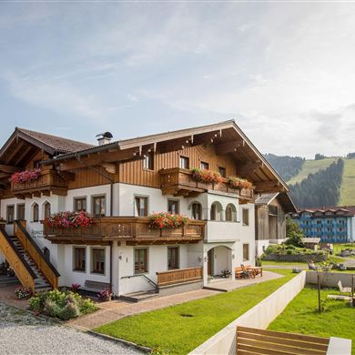 Ein gemütliches, zweigeschossiges Haus im alpinen Stil mit Balkonen und Blumen. Im Hintergrund sind grüne Hügel und ein klarer Himmel sichtbar.