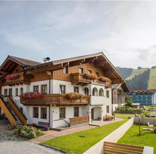 Ein gemütliches, zweigeschossiges Haus im alpinen Stil mit Balkonen und Blumen. Im Hintergrund sind grüne Hügel und ein klarer Himmel sichtbar.