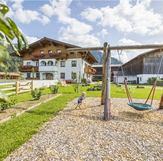 Ein schöner Spielplatz mit einer Schaukel und einer grünen Wiese. Im Hintergrund ist ein großes Haus und die Berge sind zu sehen.