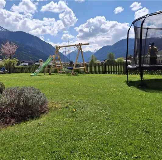 Ein schöner Garten mit einer Wiese, einem Spielplatz und einer Trampolin. Im Hintergrund sind Berge und ein blauer Himmel mit Wolken zu sehen.