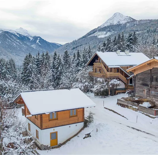 Eine winterliche Landschaft mit verschneiten Hütten und hohen Bergen im Hintergrund. Die Bäume sind mit Schnee bedeckt, was eine ruhige und idyllische Atmosphäre schafft.