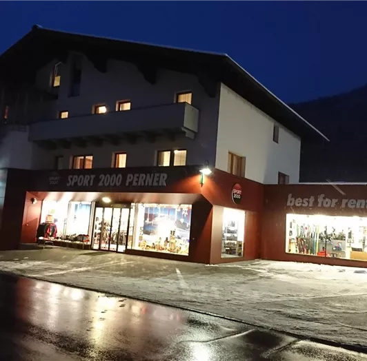 Ein modernes Geschäftshaus bei Nacht, beleuchtet von Innen. Der Außenbereich zeigt Schaufenster mit Sport- und Mietartikeln.