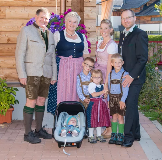 Eine fröhliche Familie in traditioneller Tracht steht vor einem Holzhaus. Im Vordergrund sitzt ein Baby in einer Tragetasche.