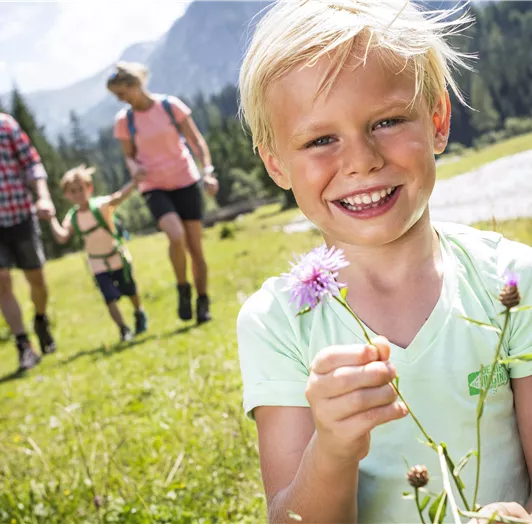 Ein fröhlicher Junge hält eine Blume in der Hand und lächelt. Im Hintergrund ist eine Familie beim Wandern zu sehen, umgeben von einer schönen Landschaft.