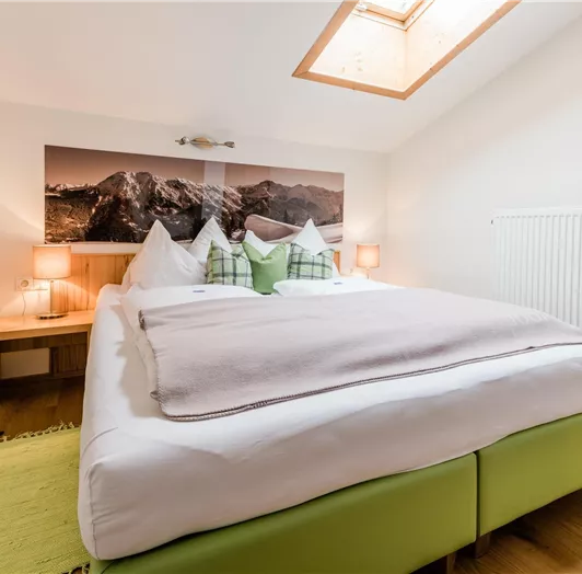 Ein gemütliches Schlafzimmer mit einem großen Bett und wolkenweißen Bettwäsche. An der Wand hängt ein Bild von Bergen, und es gibt sanfte Beleuchtung.