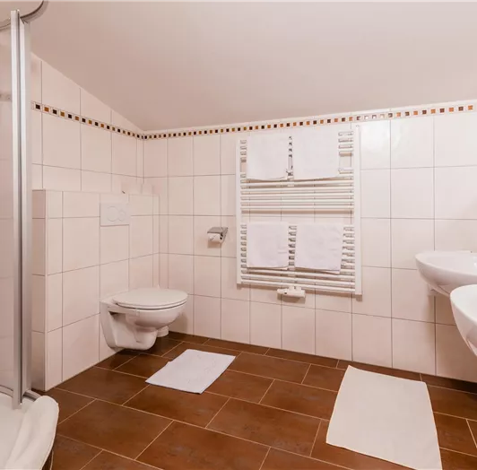 Ein modernes Badezimmer mit einer Dusche, zwei Waschbecken und einem WC. Helle Fliesen und braune Bodenfliesen sorgen für eine freundliche Atmosphäre.