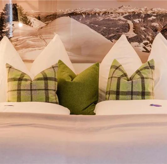 Ein gemütliches Bett mit großen, weißen Kissen und grünen Dekokissen. Im Hintergrund ist eine Berglandschaft zu sehen.