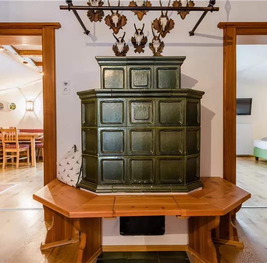 Ein traditioneller Ofen aus grünem Keramik steht im Mittelpunkt des Raumes. Die Umgebung zeigt Holzverkleidung und Zugang zu zwei anderen Räumen.