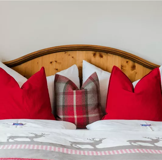 Ein gemütliches Bett mit roten Kissen und einer weißen Decke mit Rentier-Muster. Der Hintergrund ist aus Holz, was eine warme Atmosphäre schafft.