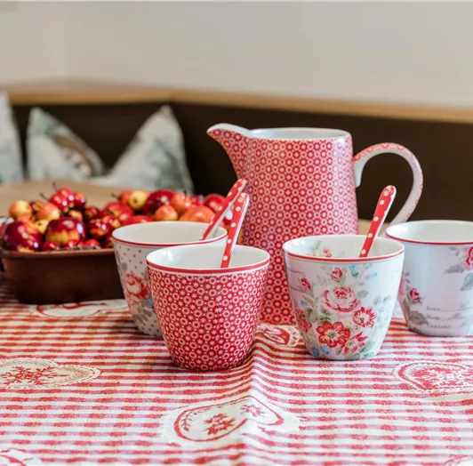 Eine einladende Tischdekoration mit roten und weißen Tassen, einer Kanne und einem Obstkorb. Der Tisch ist mit einem karierten Tischcloth gedeckt.