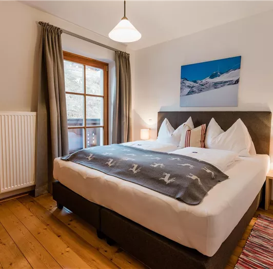 Ein gemütliches Schlafzimmer mit einem großen Bett und Holzfußboden. Die Fenster bieten einen Blick nach draußen, und es gibt warmes Licht von den Lampen.