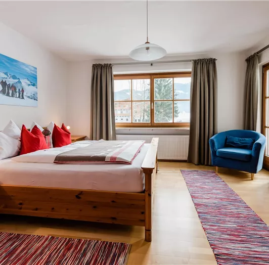 Ein gemütliches Zimmer mit einem großen Bett und roten Kissen. Es enthält einen blauen Sessel, einen Tisch und ein Fenster mit Aussicht.