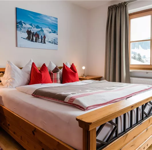 Ein gemütliches Schlafzimmer mit einem großen Holzbett und roten Kissen. Neben dem Bett stehen Nachttische, und ein Fenster bietet Blick auf die Landschaft.