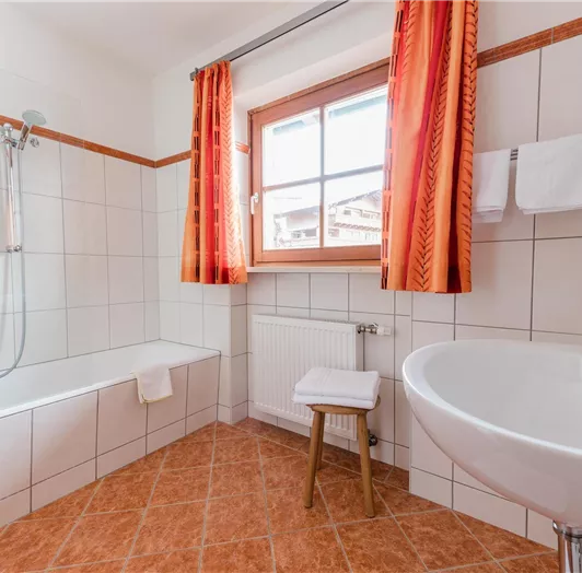 Ein helles Badezimmer mit einer Badewanne und einer Dusche. Die Fenster sind mit roten Vorhängen versehen, und es gibt ein Waschbecken sowie Handtücher.