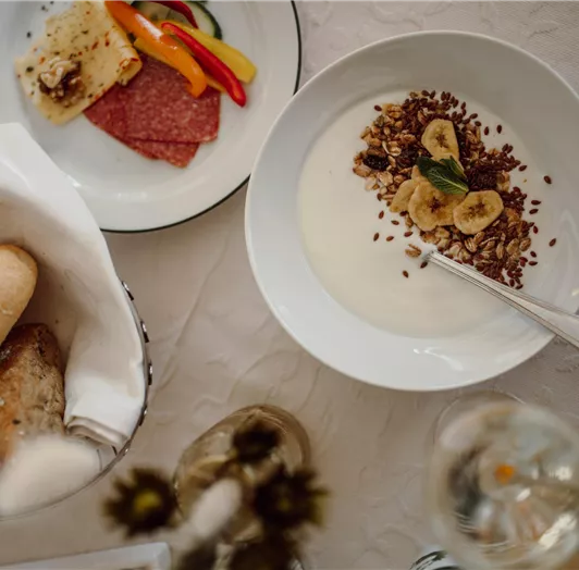 A beautiful breakfast table with yogurt, muesli, and bananas. There are also cold cuts, vegetables, and rolls.