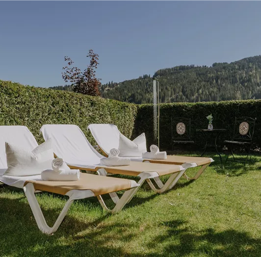 Eine rustikale Gartenlandschaft mit Liegestühlen auf grünem Rasen. Im Hintergrund sind Hügel und eine dichte Hecke zu sehen.