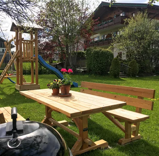 Ein großzügiger Garten mit einem Holztisch und Bänken. Im Hintergrund ist ein Spielplatz mit einer Rutsche zu sehen.