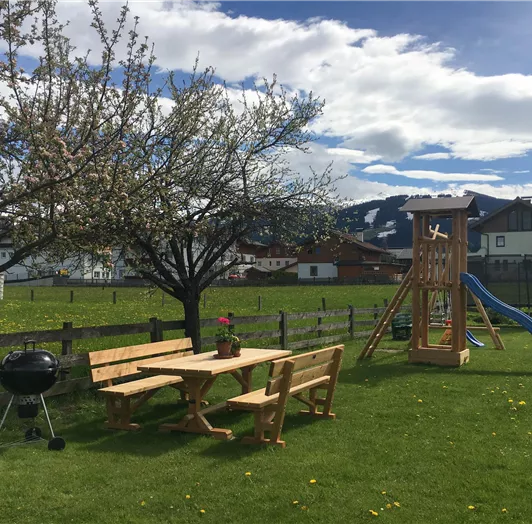 Ein schöner Garten mit einem Holztisch, Bänken und einem Grill. Im Hintergrund steht ein Spielgerät mit Rutsche und eine blühende Baumreihe.