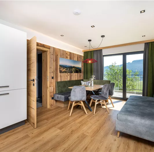 Ein modernes Wohnzimmer mit Holz- und Grüntönen. Es gibt einen Esstisch, einen bequemen Sitzbereich und große Fenster mit Blick auf die Berge.