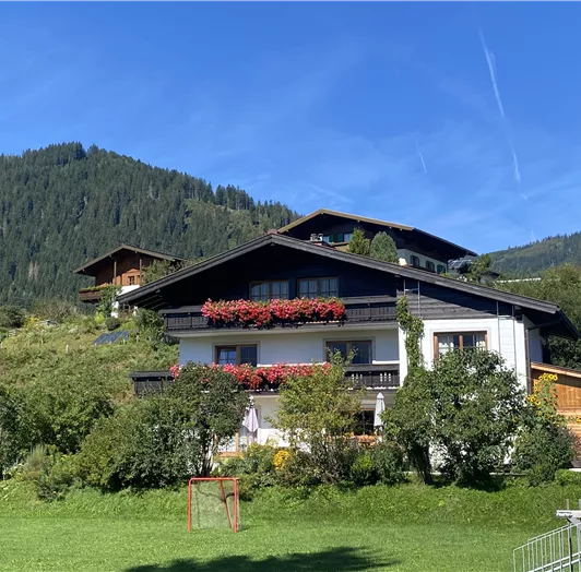 Ein malerisches Haus in den Bergen mit bunten Blumenbalkonen. Umgeben von einer grünen Wiese und sanften Hügeln.