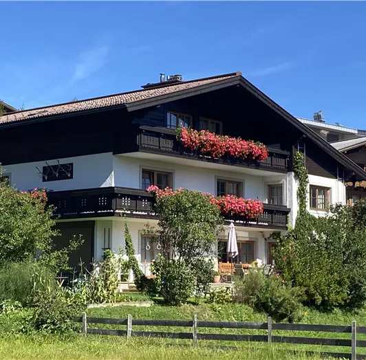 Ein traditionelles alpenländisches Haus mit bunten Blumen auf dem Balkon. Umgeben von Grünflächen und Bäumen unter einem blauen Himmel.