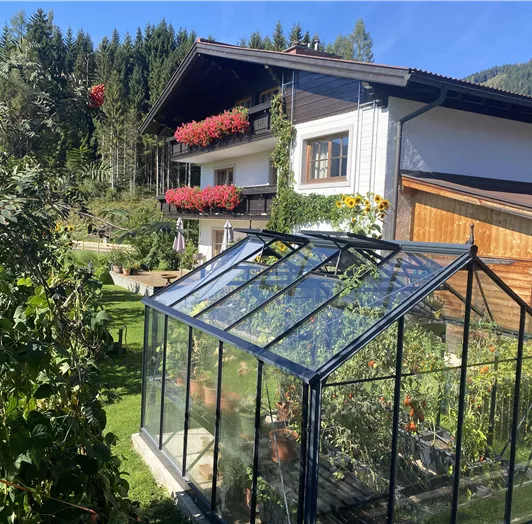 Ein grüner Garten mit einem Gewächshaus und blühenden Pflanzen. Im Hintergrund steht ein Haus mit bunten Balkonblumen.