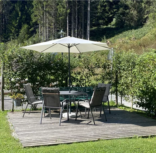 Ein gemütlicher Tisch mit Stühlen unter einem Sonnenschirm steht auf einer Holzer Terrasse. Umgeben von grünen Pflanzen und Wald.