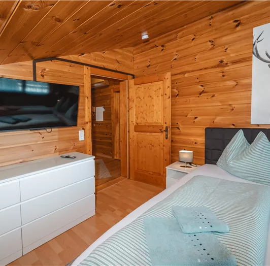 Ein gemütliches Schlafzimmer mit Holzwänden und einem modernen Fernseher. Das Bett ist mit traditionellen Kissen in sanften Farben dekoriert.
