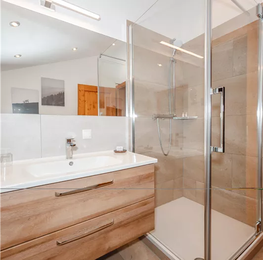 Ein modernes Badezimmer mit einer begehbaren Dusche aus Glas und einem Holzschrank. Die Beleuchtung ist hell und schafft eine einladende Atmosphäre.