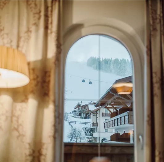 A view from a window onto a snowy landscape with ski lifts in Flachau. In the foreground, curtains and a lamp are visible - direct view from the room window.