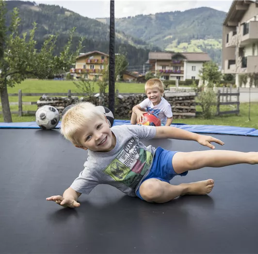 Zwei Kinder springen auf einem Trampolin. Sie haben Spaß in einer grünen Landschaft mit Bergen im Hintergrund.