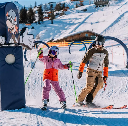 Ein Kind in lila Schneeanzug und ein Erwachsener in braunem Anzug fahren Ski auf einer verschneiten Piste. Im Hintergrund sind ein Skilift und ein Holzhaus sichtbar.