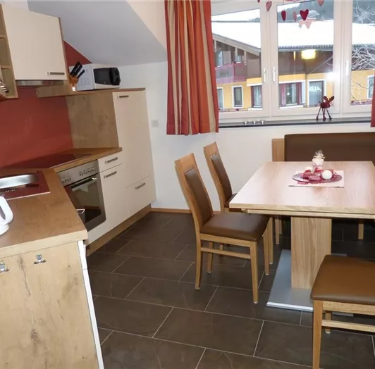 Eine moderne Küche mit Holzmöbeln und einem Esstisch. Der Raum ist hell und freundlich, mit großen Fenstern, die Blick nach draußen geben.