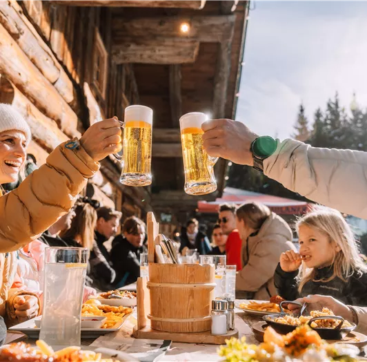 Eine Gruppe von Menschen sitzt an einem Tisch in der Sonne und hebt Biergläser zum anstoßen. Hinter ihnen sind viele Gäste und eine gemütliche Hütte zu sehen.
