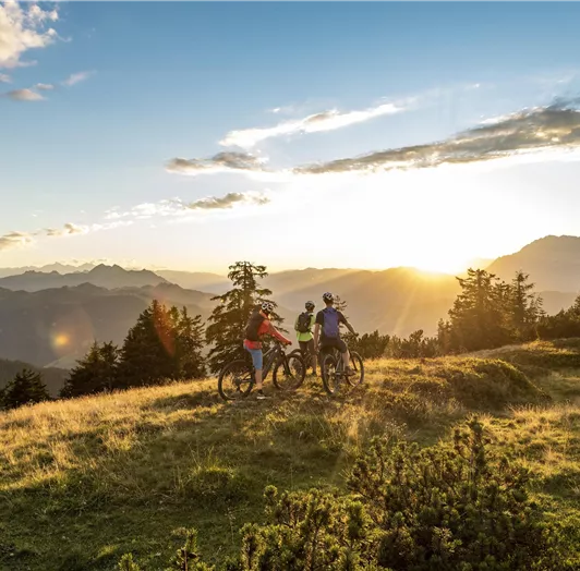 Zwei Radfahrer genießen den Sonnenuntergang auf einem Hügel mit Blick auf die Berge. Die Landschaft ist grün und üppig mit Baumgruppen.
