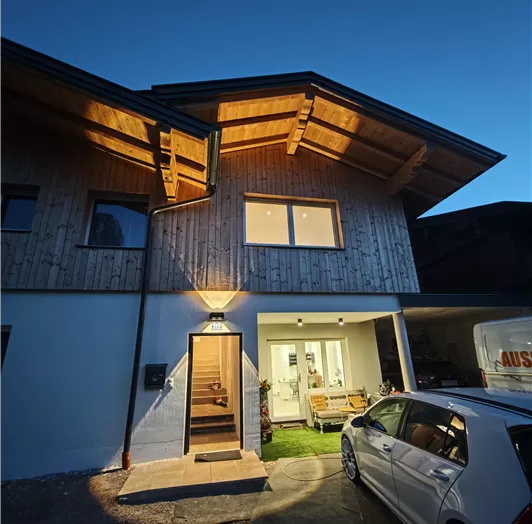 Ein modernes Haus mit Holzelementen und einem großen Walmdach.  Die Abendbeleuchtung sorgt für eine gemütliche Atmosphäre.