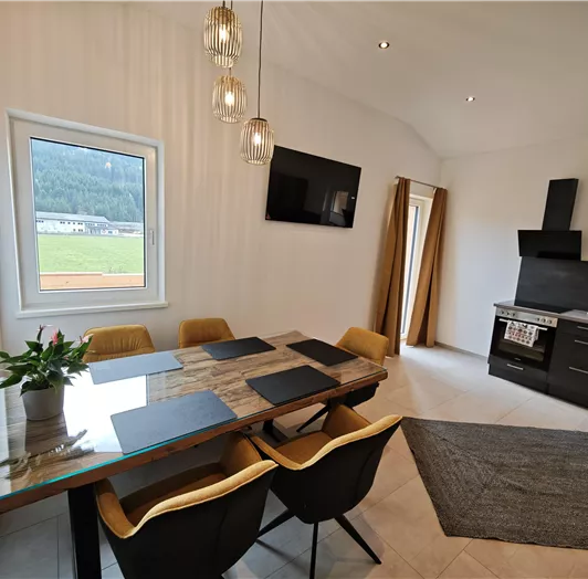 Ein modernes Esszimmer mit einem großen Tisch, Stühlen und einem Fenster, das auf eine grüne Landschaft blickt. An der Wand befindet sich ein Fernseher und eine moderne Küchenzeile.