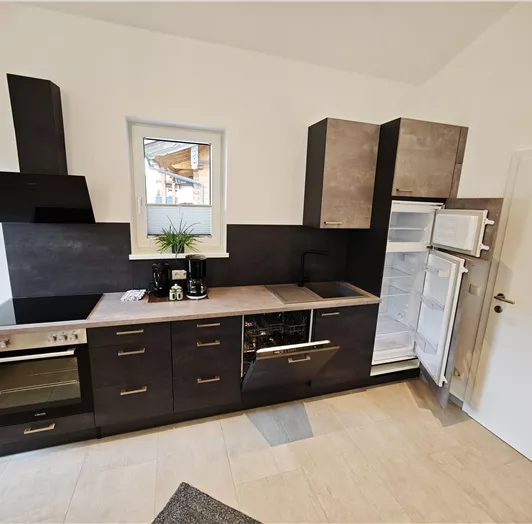 Eine moderne Küche mit dunklen Holzschränken und einer hellen Arbeitsfläche. Sie verfügt über einen Kühlschrank, einen Herd und eine Spüle, sowie ein Fenster, das Tageslicht hereinlässt.